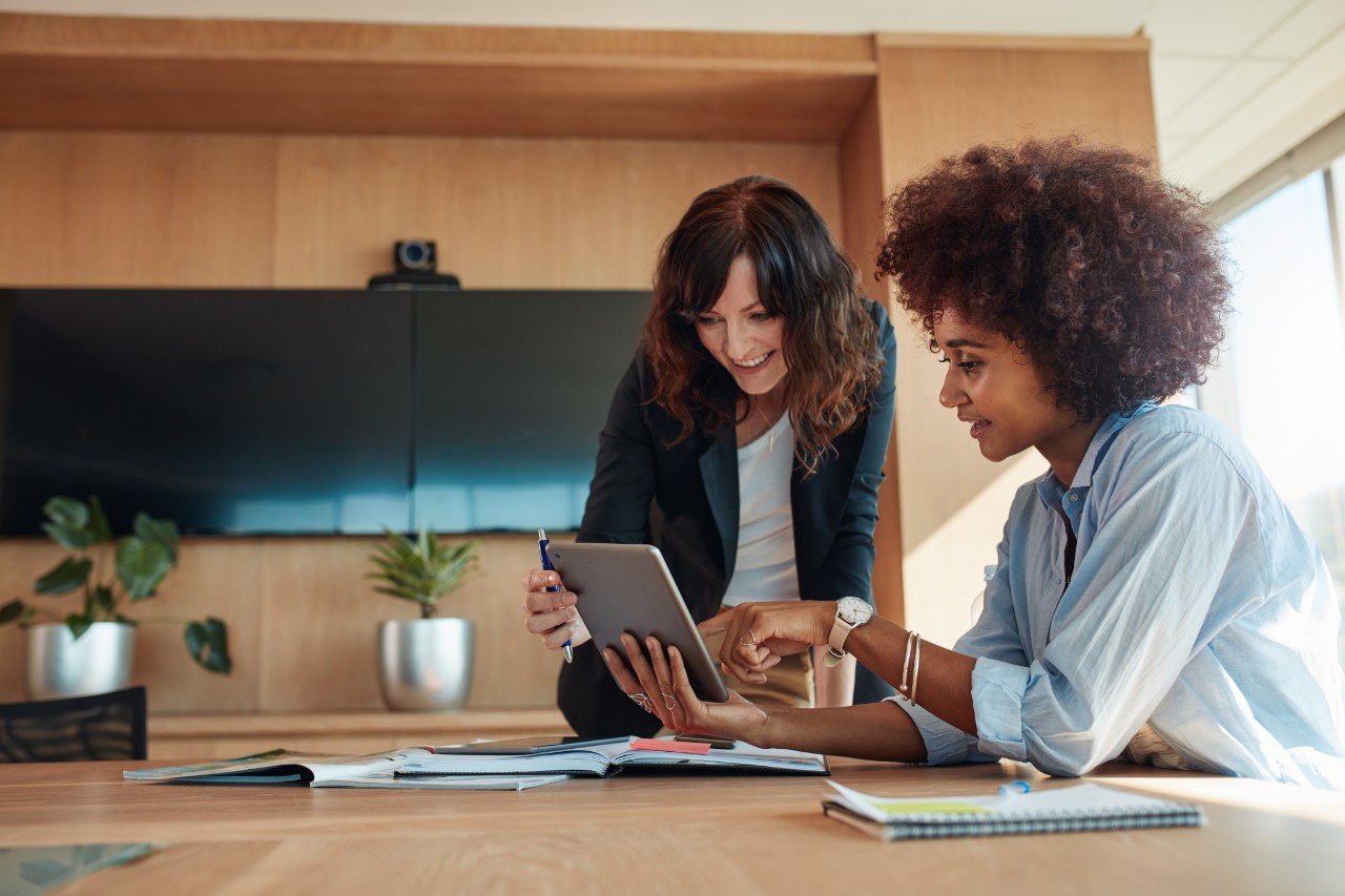 African businesswoman showing something on digital tablet to her female colleague while sitting at her desk. Professional employees discussing ideas of project on tablet pc.